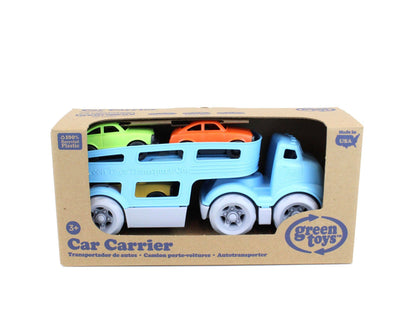 Green Toys Car Carrier in retail packaging on white background — kraft brown cardboard box with large die-cut window showing the blue car carrier truck inside with green and orange Mini Cars on the upper deck and yellow car visible in the lower level; Green Toys logo badge in lower right corner; "Car Carrier" in bold text across the bottom in English, Spanish, French, and German; "3+" age label in lower left; "100% Recycled Plastic" and "Made in USA" badges visible on the box