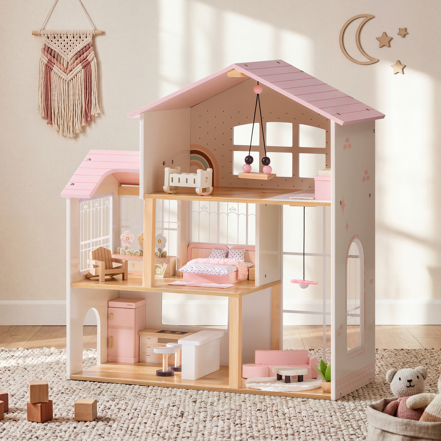 Robud 3-story pink and cream wooden dollhouse in a styled boho nursery — pink macrame wall hanging above, children's room on top floor with crib and mobile, bedroom on middle floor with pink bedding, kitchen and living room on ground floor; cream shag rug and woven basket with teddy bear beside it