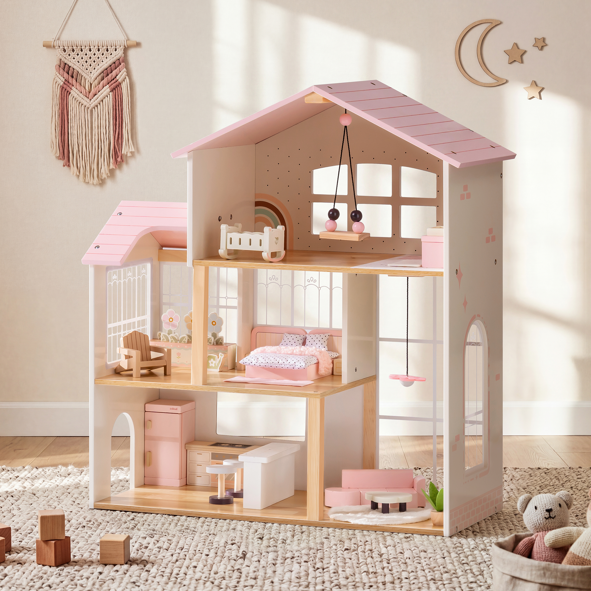 Robud 3-story pink and cream wooden dollhouse in a styled boho nursery — pink macrame wall hanging above, children's room on top floor with crib and mobile, bedroom on middle floor with pink bedding, kitchen and living room on ground floor; cream shag rug and woven basket with teddy bear beside it
