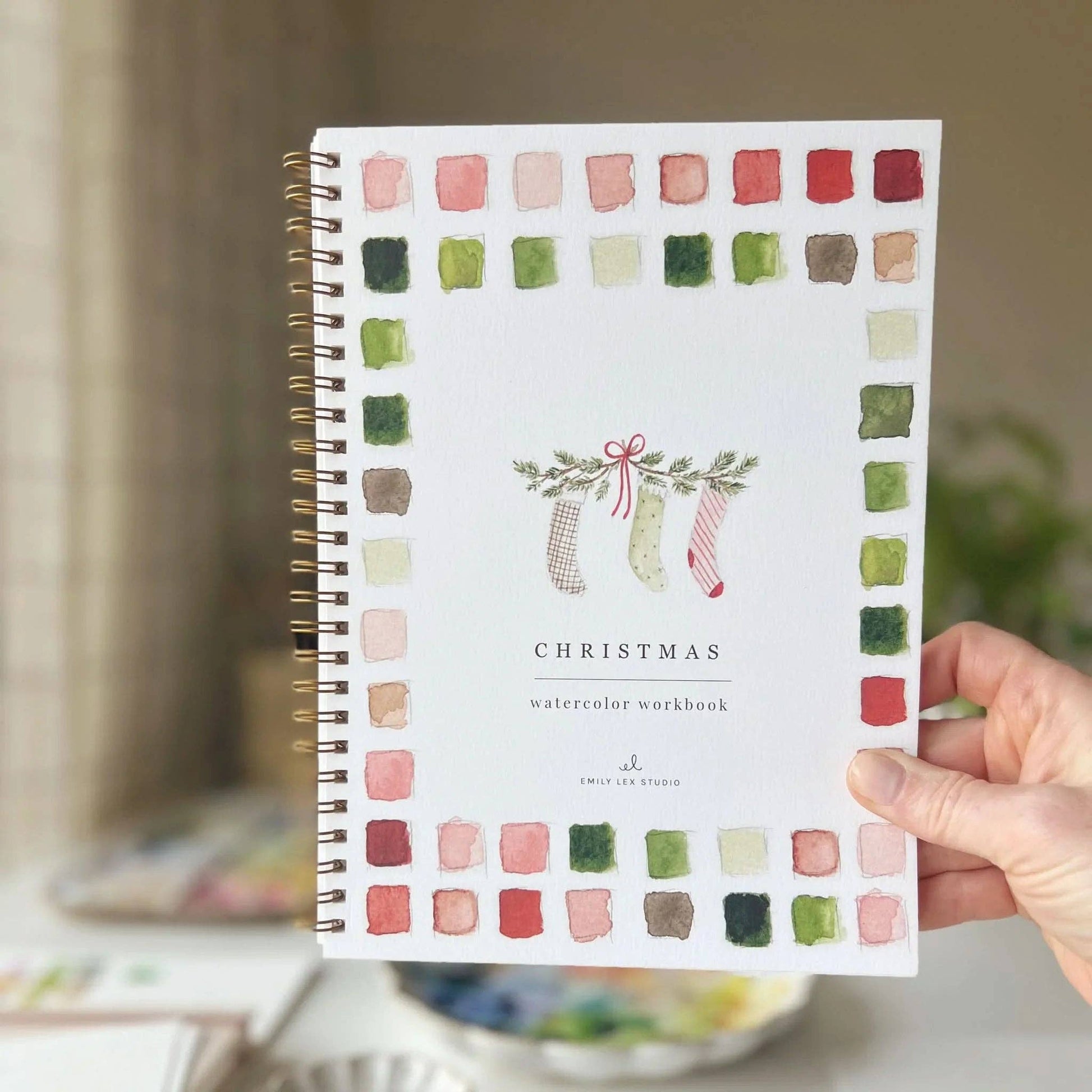 Hand holding the Christmas Watercolor Workbook by Emily Lex Studio — cover showing three Christmas stockings hanging from a pine garland with a red bow, bordered by red, green, and pink watercolor swatches
