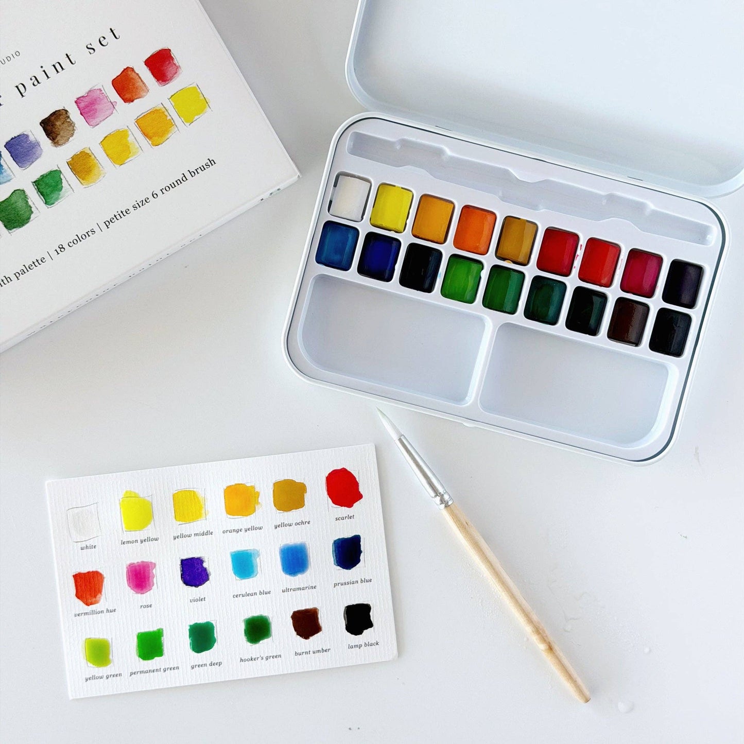 Emily Lex Studio watercolor paint set flat lay on white surface; open tin at upper right showing 18 paint pans in three rows with two mixing trays at bottom; product box at upper left; filled-out color swatch card at lower center with all 18 colors painted in; size 6 round brush with wood handle resting diagonally across the swatch card


