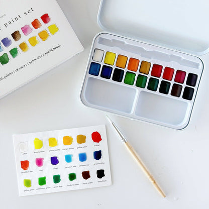 Emily Lex Studio watercolor paint set flat lay on white surface; open tin at upper right showing 18 paint pans in three rows with two mixing trays at bottom; product box at upper left; filled-out color swatch card at lower center with all 18 colors painted in; size 6 round brush with wood handle resting diagonally across the swatch card


