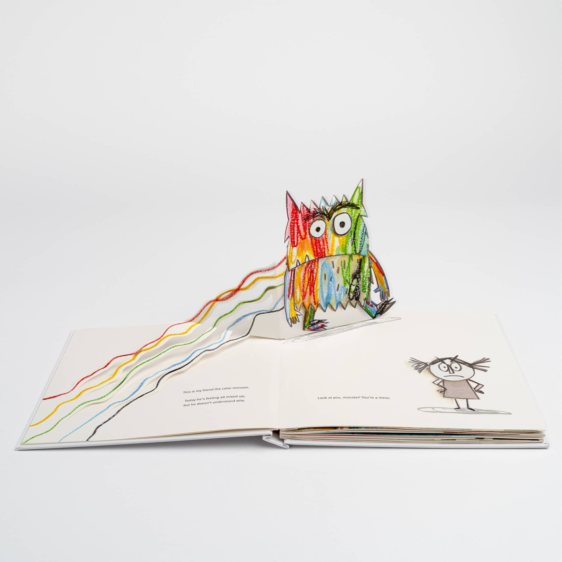 Open spread of The Color Monster: A Pop-Up Book of Feelings showing the opening page — 3-D pop-up of the multicolor Color Monster standing amid tangled emotion lines in yellow, red, green, blue, and black, with a sketched girl on the right saying "Look at you, monster! You're a mess," on a white background
