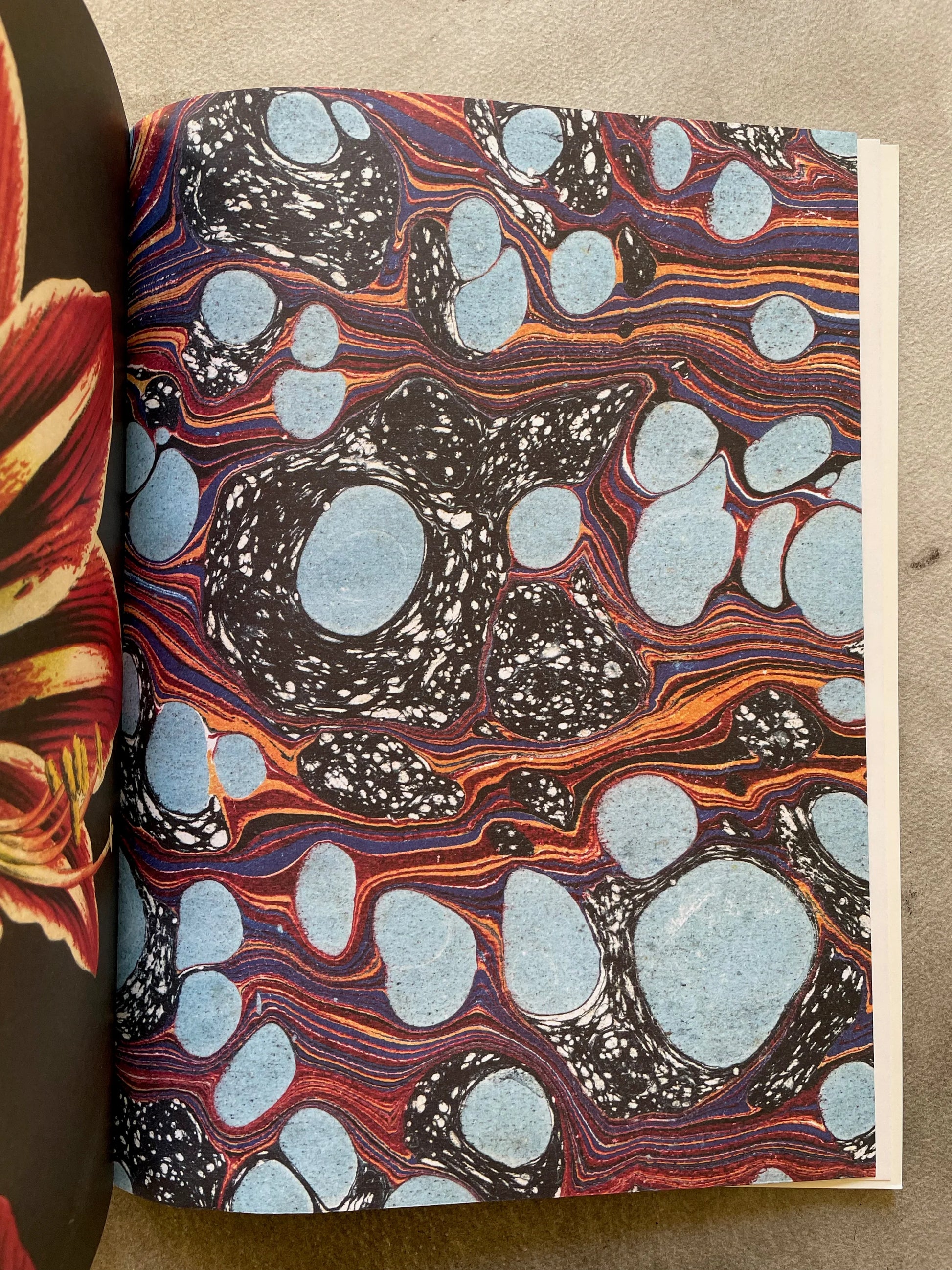 Open spread of John Derian Wrapping Paper & Gift Tags showing the marbled wrapping paper sheet — bold blue oval shapes on a swirling black, red, orange, and purple marbled background, with a glimpse of the red amaryllis sheet on the left, photographed on a gray surface