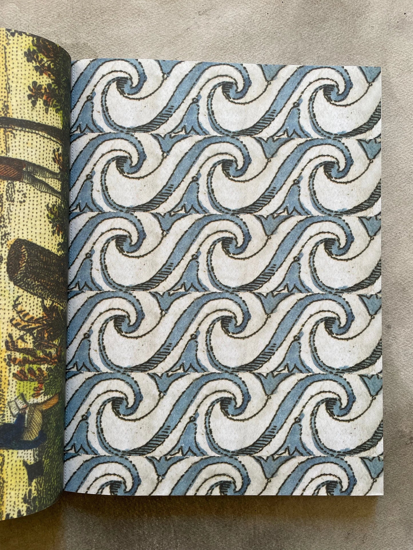 Open spread of John Derian Wrapping Paper & Gift Tags showing the blue scrollwork ocean wave wrapping paper sheet — repeating interlocking blue and white spiral wave pattern sourced from 19th-century printed matter, with a glimpse of a naturalist illustration sheet on the left, photographed on a gray surface