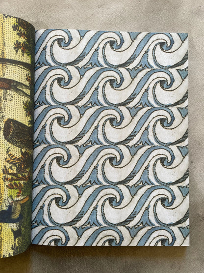 Open spread of John Derian Wrapping Paper & Gift Tags showing the blue scrollwork ocean wave wrapping paper sheet — repeating interlocking blue and white spiral wave pattern sourced from 19th-century printed matter, with a glimpse of a naturalist illustration sheet on the left, photographed on a gray surface