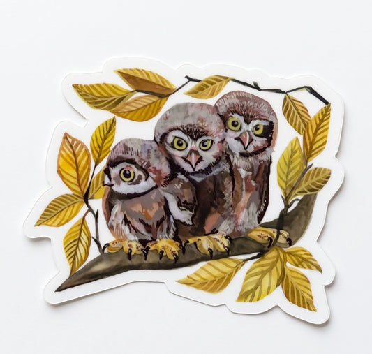 Made by Shellie Owl Trio vinyl sticker — three wide-eyed baby owlets with yellow-green eyes perched together on a branch surrounded by golden autumn leaves in warm browns and yellows; die-cut with white border; white background