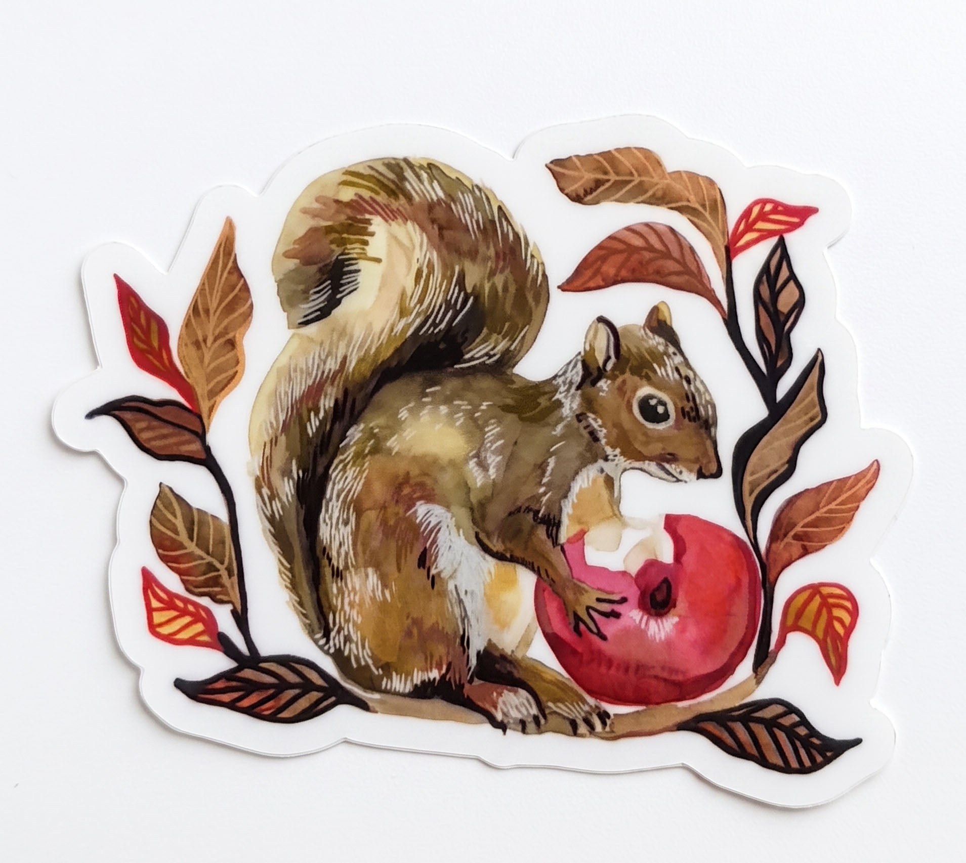 Made by Shellie Squirrel vinyl sticker — detailed illustrated squirrel clutching a red apple surrounded by autumn leaves in rich reds, oranges, and browns; die-cut with white border; white background